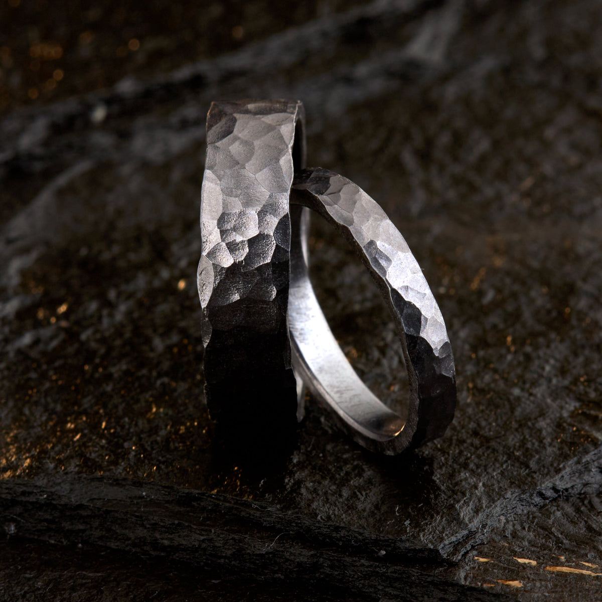 Handgefertigtes Ehering-Set aus massivem Tantal auf rustikalem Leder. Herrenring (6mm) und Damenring (4mm) mit markanter, gehämmerter Oberfläche in dunklem Anthrazit. Einzigartige Schmiedekunst für exklusive Hochzeitsringe. SEO-Keywords: Tantal Eheringe gehämmert, dunkle Trauringe, Tantalum Wedding Bands, nachhaltiger Luxusschmuck, seltene Edelmetalle Hochzeit. Sichtbare Details der individuellen Textur und des tiefen, metallischen Glanzes.