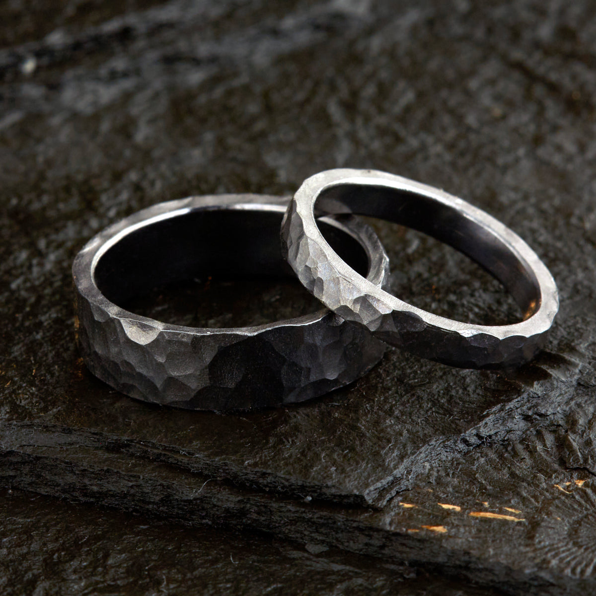 Handgefertigtes Ehering-Set aus massivem Tantal auf rustikalem Leder. Herrenring (6mm) und Damenring (4mm) mit markanter, gehämmerter Oberfläche in dunklem Anthrazit. Einzigartige Schmiedekunst für exklusive Hochzeitsringe. SEO-Keywords: Tantal Eheringe gehämmert, dunkle Trauringe, Tantalum Wedding Bands, nachhaltiger Luxusschmuck, seltene Edelmetalle Hochzeit. Sichtbare Details der individuellen Textur und des tiefen, metallischen Glanzes.