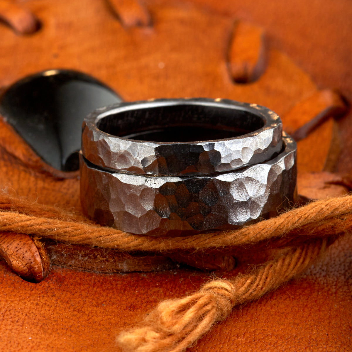 Handgefertigtes Ehering-Set aus massivem Tantal auf rustikalem Leder. Herrenring (6mm) und Damenring (4mm) mit markanter, gehämmerter Oberfläche in dunklem Anthrazit. Einzigartige Schmiedekunst für exklusive Hochzeitsringe. SEO-Keywords: Tantal Eheringe gehämmert, dunkle Trauringe, Tantalum Wedding Bands, nachhaltiger Luxusschmuck, seltene Edelmetalle Hochzeit. Sichtbare Details der individuellen Textur und des tiefen, metallischen Glanzes.