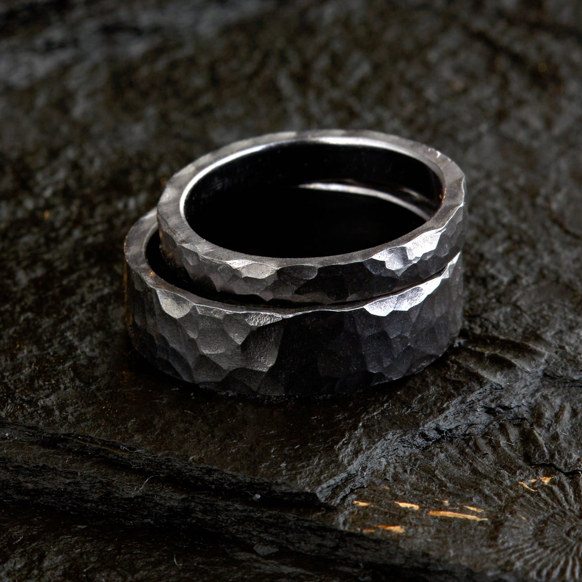 Handgefertigtes Ehering-Set aus massivem Tantal auf rustikalem Leder. Herrenring (6mm) und Damenring (4mm) mit markanter, gehämmerter Oberfläche in dunklem Anthrazit. Einzigartige Schmiedekunst für exklusive Hochzeitsringe. SEO-Keywords: Tantal Eheringe gehämmert, dunkle Trauringe, Tantalum Wedding Bands, nachhaltiger Luxusschmuck, seltene Edelmetalle Hochzeit. Sichtbare Details der individuellen Textur und des tiefen, metallischen Glanzes.