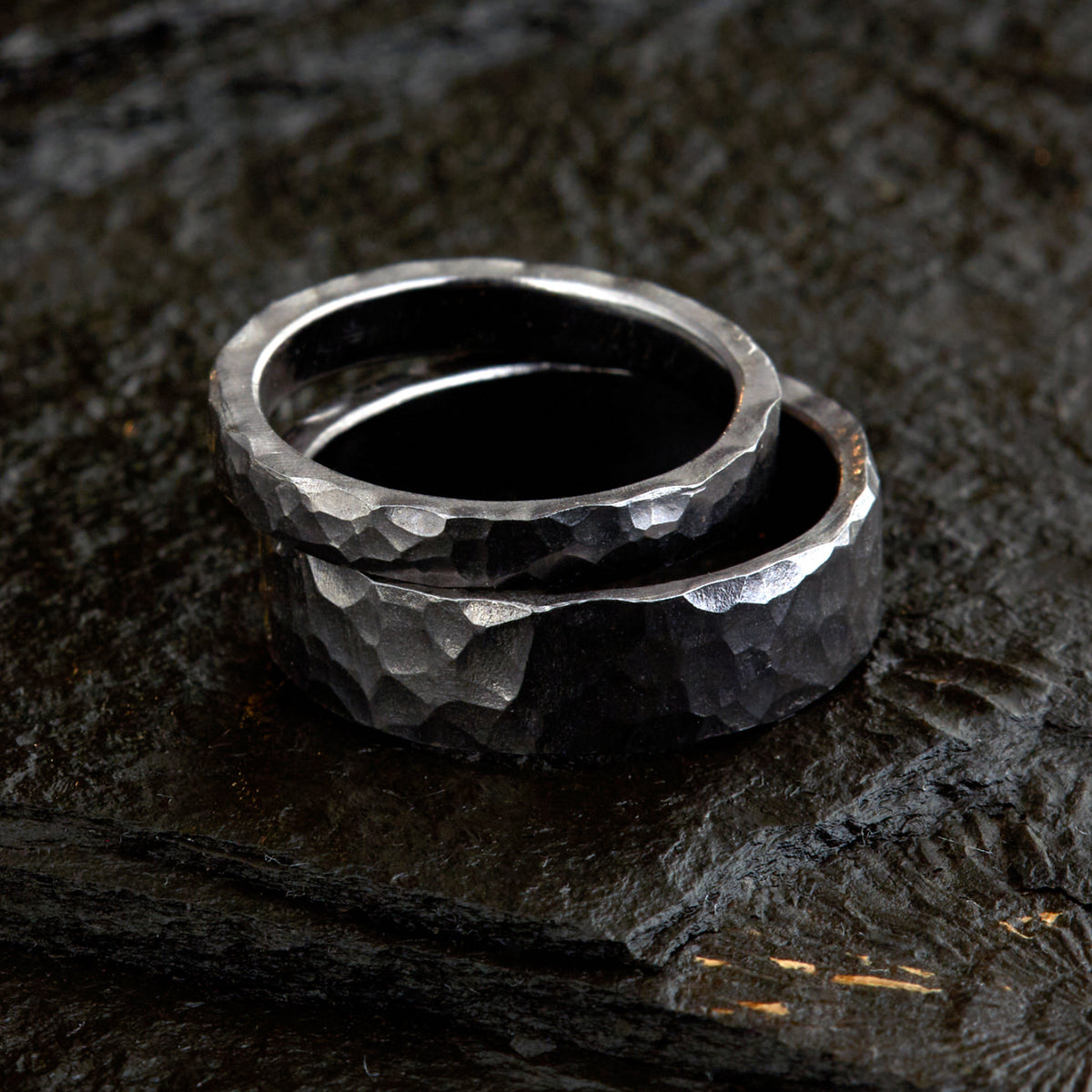Handgefertigtes Ehering-Set aus massivem Tantal auf rustikalem Leder. Herrenring (6mm) und Damenring (4mm) mit markanter, gehämmerter Oberfläche in dunklem Anthrazit. Einzigartige Schmiedekunst für exklusive Hochzeitsringe. SEO-Keywords: Tantal Eheringe gehämmert, dunkle Trauringe, Tantalum Wedding Bands, nachhaltiger Luxusschmuck, seltene Edelmetalle Hochzeit. Sichtbare Details der individuellen Textur und des tiefen, metallischen Glanzes.