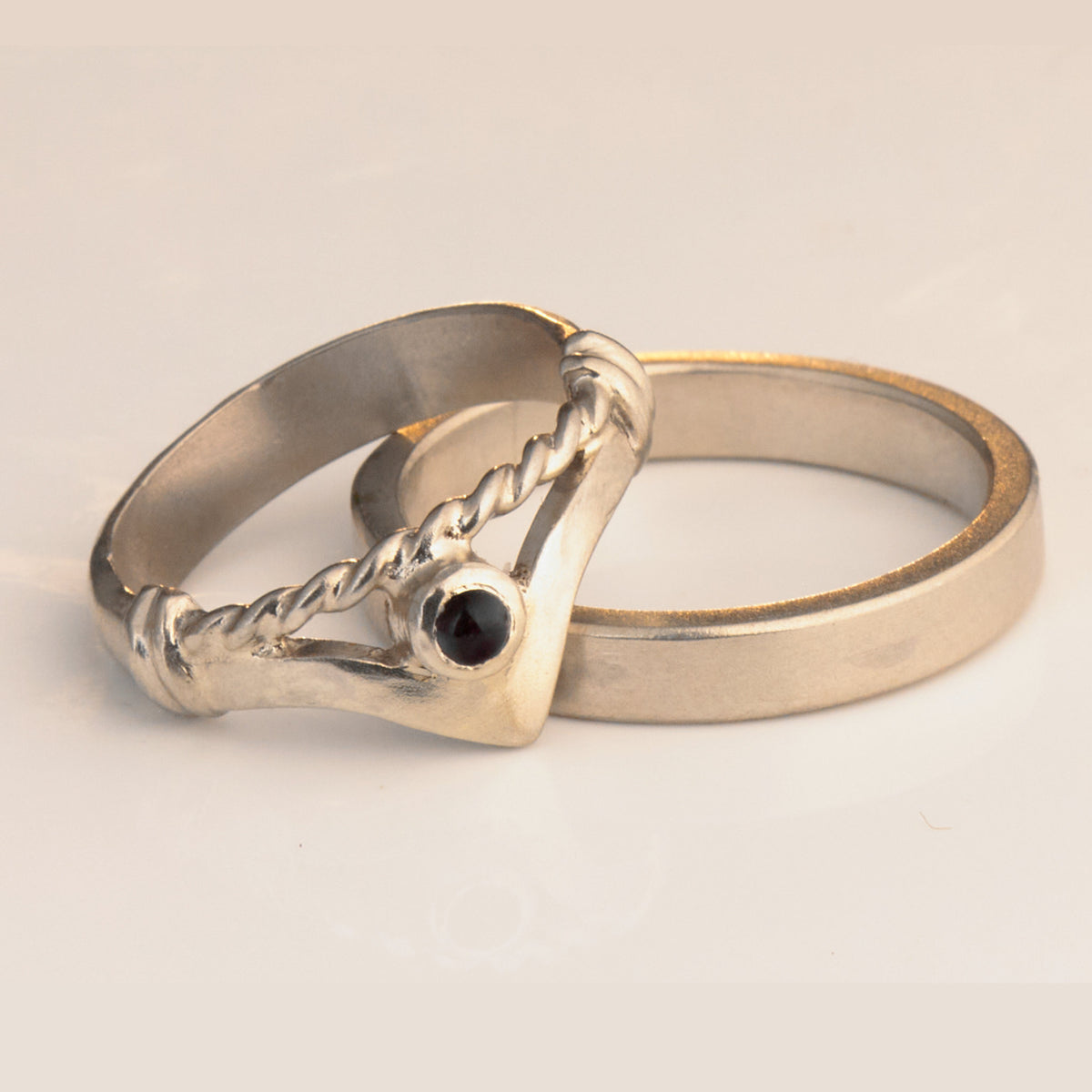 Handgefertigter Vintage Wishbone-Ring aus mattem 925 Silber mit einem violetten 3 mm Amethyst Cabochon in Zargenfassung. Man sieht deutlich die geteilte Schiene mit der filigranen Kordel-Verblendung.
Set-Präsentation: Minimalistisches Ehering-Set von Silberschweif auf dunklem Stein. Der verspielte Damenring mit lila Edelstein harmoniert perfekt mit dem schlichten, 4 mm breiten matten Herrenring.
Detail Ringschiene: Seitenansicht des Damenrings zeigt die handwerkliche Aufteilung der 4 mm Silberschiene 