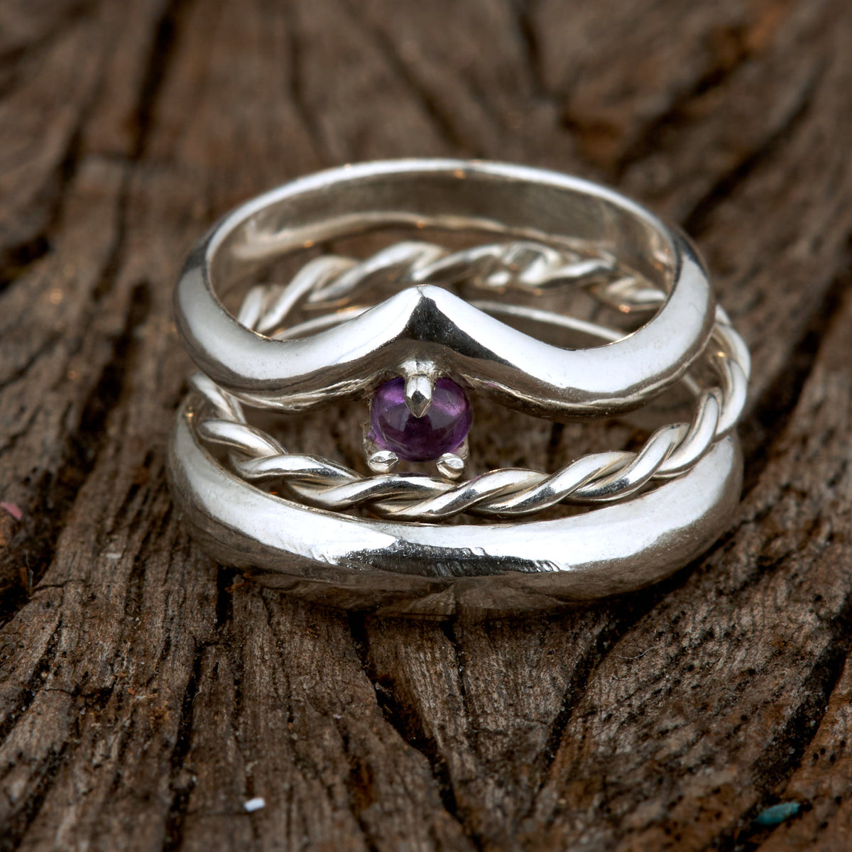 Handgefertigtes Ehering-Set aus 925 Silber auf Leder. 3-teiliges Damen-Set bestehend aus einem 3mm Wishbone-Ring mit Amethyst und einem 2mm Kordelring. Daneben ein massiver 4mm Herrenring im Halbrund-Profil. Zeitloses Design mit lila Edelstein für die Hochzeit. SEO-Keywords: Wishbone Eheringe Silber, Amethyst Trauringe Set, handgefertigte Silberringe, 925 Sterlingsilber Schmuck, Kordelring Hochzeit. Sichtbare Details der hochwertigen Handarbeit und Steinbesetzung.