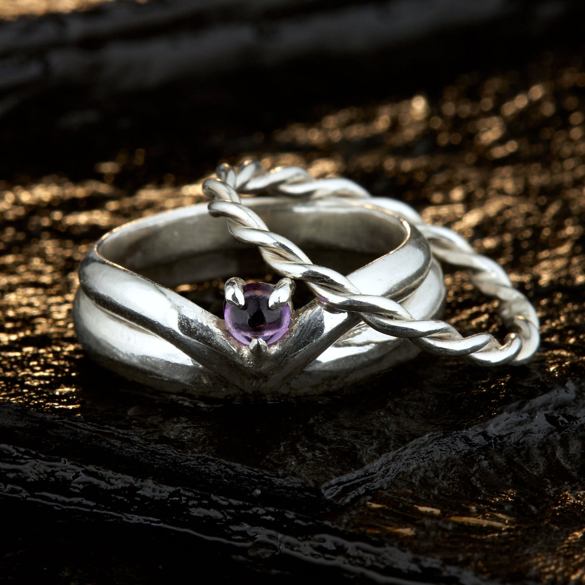 Handgefertigtes Ehering-Set aus 925 Silber auf Leder. 3-teiliges Damen-Set bestehend aus einem 3mm Wishbone-Ring mit Amethyst und einem 2mm Kordelring. Daneben ein massiver 4mm Herrenring im Halbrund-Profil. Zeitloses Design mit lila Edelstein für die Hochzeit. SEO-Keywords: Wishbone Eheringe Silber, Amethyst Trauringe Set, handgefertigte Silberringe, 925 Sterlingsilber Schmuck, Kordelring Hochzeit. Sichtbare Details der hochwertigen Handarbeit und Steinbesetzung.