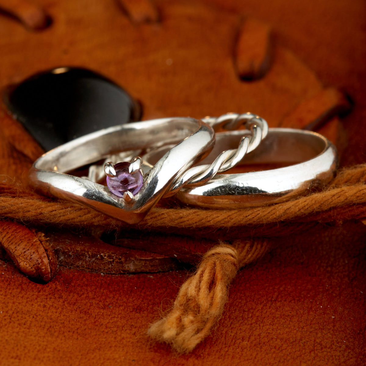 Handgefertigtes Ehering-Set aus 925 Silber auf Leder. 3-teiliges Damen-Set bestehend aus einem 3mm Wishbone-Ring mit Amethyst und einem 2mm Kordelring. Daneben ein massiver 4mm Herrenring im Halbrund-Profil. Zeitloses Design mit lila Edelstein für die Hochzeit. SEO-Keywords: Wishbone Eheringe Silber, Amethyst Trauringe Set, handgefertigte Silberringe, 925 Sterlingsilber Schmuck, Kordelring Hochzeit. Sichtbare Details der hochwertigen Handarbeit und Steinbesetzung.