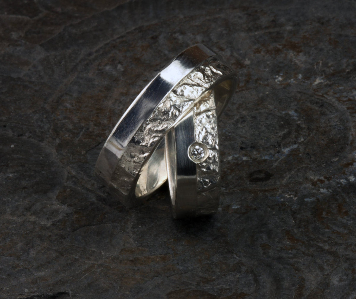 Massive Trauringe aus 925 Silber handgefertigt. Eheringe mit markanter Schieferstruktur retikuliert im Kontrast zu mattierten Oberflächen. Damenring ca. 6mm breit mit einem bündig gefassten 2mm Brillanten (TW/si). Herrenring ca. 6mm breit, massiv und charakterstark. Einzigartiges Partnerring-Set mit organischer Textur, inspiriert von natürlichem Fels. Handgemacht in eigener Werkstatt in Deutschland, keine CNC-Massenware. Hochwertige Unikate für Individualisten
